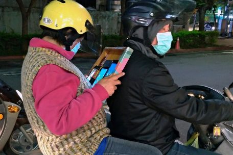 Eine Frau mit Mundschutz sitzt in Taiwan als Beifahrerin eines Mannes, der ebenfalls einen Mundschutz tr&auml;gt, auf einem Motorrad und liest auf einem Smartphone und einem Tablett.