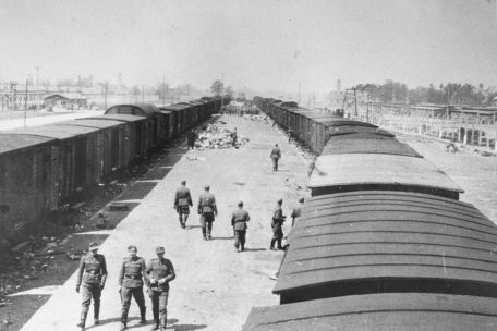 SS-M&auml;nner gehen entlang der neuen Ankunftsrampe in Auschwitz-Birkenau. Die Rampe wurde verl&auml;ngert, damit die Todesz&uuml;ge ab Fr&uuml;hjahr 1944 direkt bis zu den Vernichtungsanlagen heranfahren konnten. Die Krematorien II und III (erkennbar an den Schornsteinen) sind im Hintergrund rechts und links von den G&uuml;terwaggons zu sehen. Foto von Bernhard Walter/Ernst Hofmann, Mai 1944.